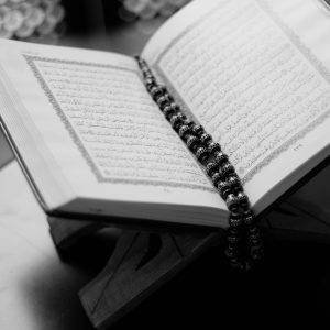 A black and white close-up of an open Quran on a wooden stand with prayer beads, symbolizing Islamic faith and devotion.